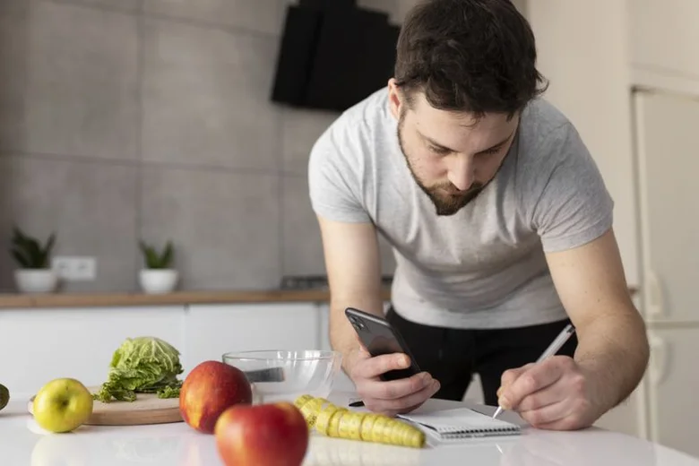 Muž počítá kalorie pomocí telefonu a poznámek v kuchyni