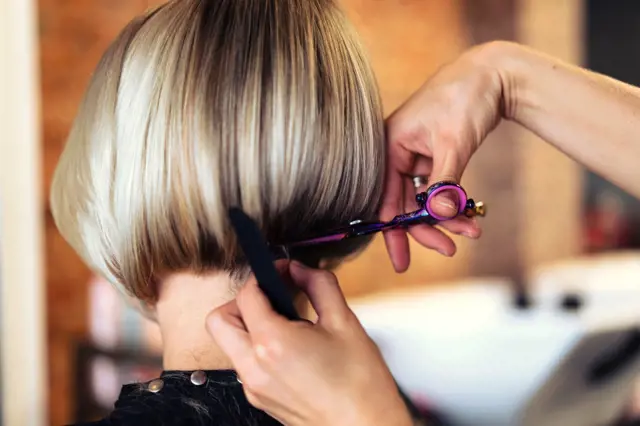 Stylista stříhá blond vlasy do podkovového střihu s ofinou.