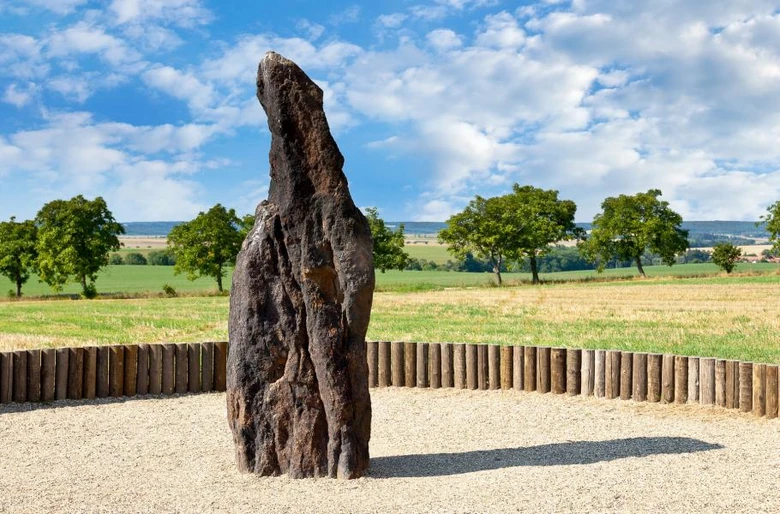 Velký kamenný menhir stojí na travnatém poli pod modrou oblohou.