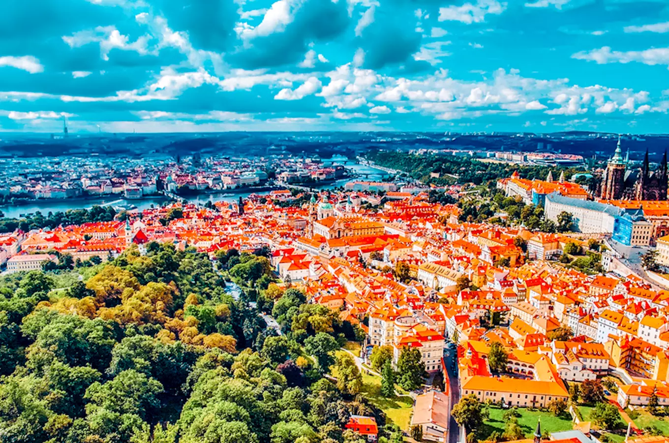 Pohled na Prahu s červenými střechami a řekou pod modrou oblohou.