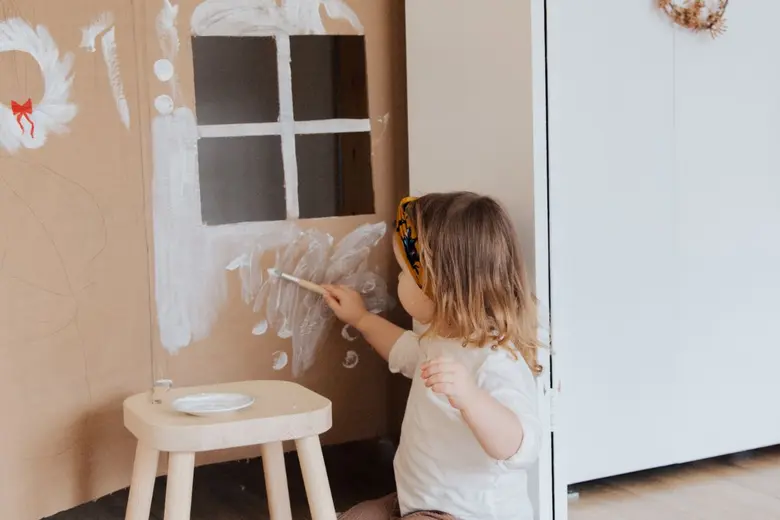 Dívka maluje bílou barvou dům z kartonu pro děti