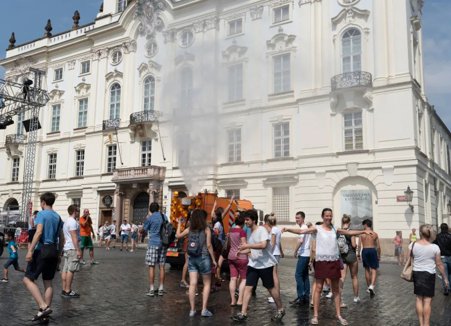 Lidé procházejí náměstím s umělým kouřem před bílou budovou.