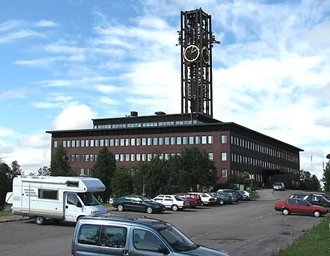 Velká budova s věží hodin v parkovišti pod modrou oblohou.
