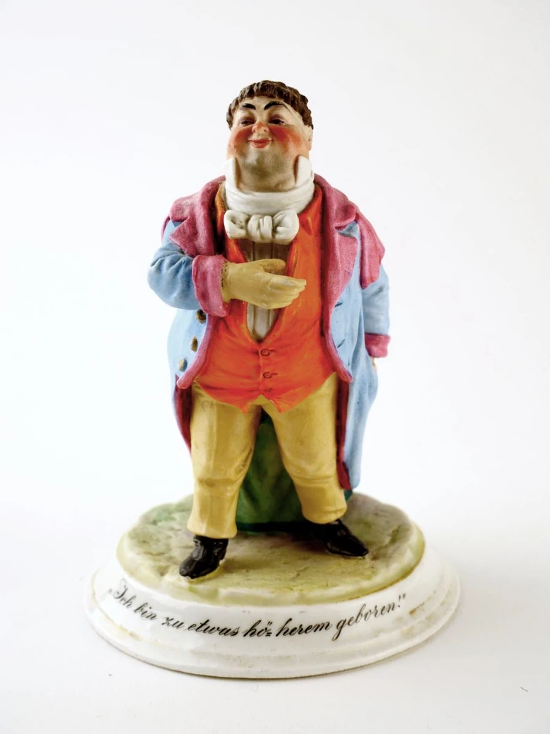 Starožitná porcelánová figurina muže na podstavci s německým nápisem.